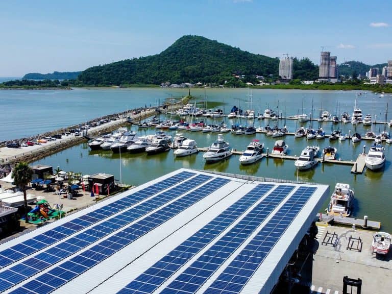 Vista aérea da Marina de Itajaí com diversos barcos atracados, painéis solares no telhado em primeiro plano e montanhas verdes ao fundo.
