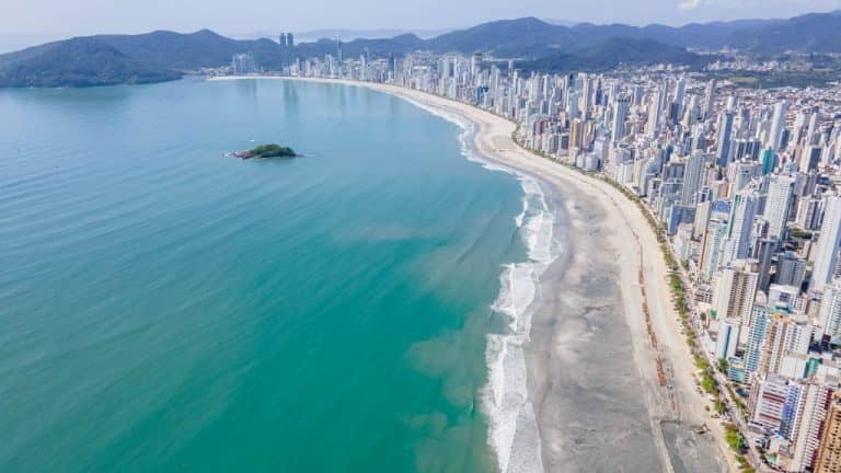 Fotografia aérea de drone da extensa faixa de areia e do mar esverdeado de Balneário Camboriú, com morros ao fundo e arranha-céus alinhados à costa.