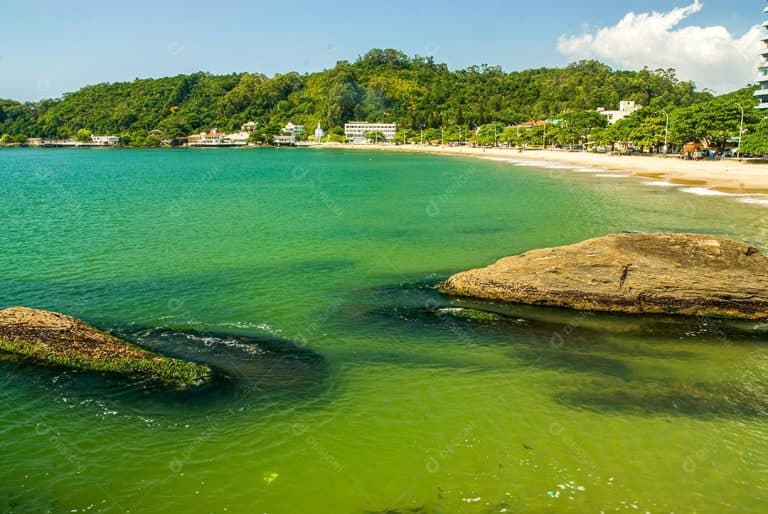 Rochas no mar de águas esverdeadas em primeiro plano, com a faixa de areia clara da Praia de Cabeçudas e o morro coberto de vegetação ao fundo.