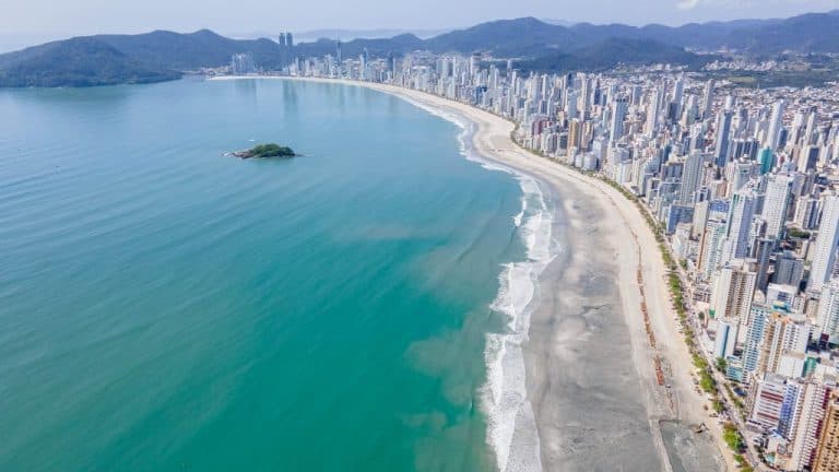 Visão aérea da orla de Balneário Camboriú, com faixa de areia, mar esverdeado e arranha-céus ao fundo.