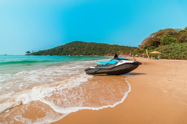 Jet ski estacionado na beira de uma praia de areia dourada, com ondas suaves, mar azul-turquesa, colinas verdes ao fundo e céu limpo.