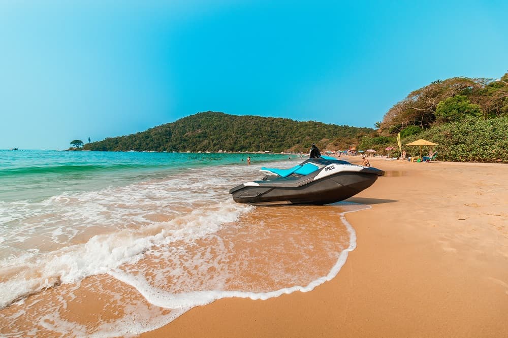 Jet ski estacionado na beira de uma praia de areia dourada, com ondas suaves, mar azul-turquesa, colinas verdes ao fundo e céu limpo.
