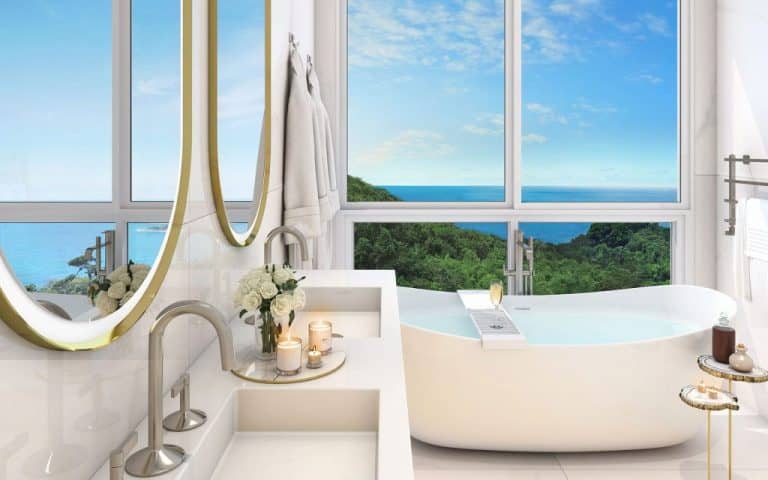 Banheiro da suíte master com bancada dupla em mármore branco, dois espelhos redondos com moldura dourada, banheira independente ao lado de janelas panorâmicas com vista para o mar e decoração com velas aromáticas.