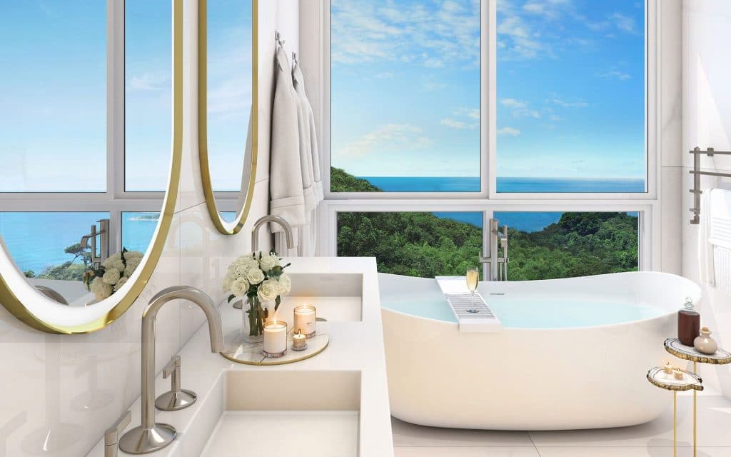 Banheiro da suíte master com bancada dupla em mármore branco, dois espelhos redondos com moldura dourada, banheira independente ao lado de janelas panorâmicas com vista para o mar e decoração com velas aromáticas.