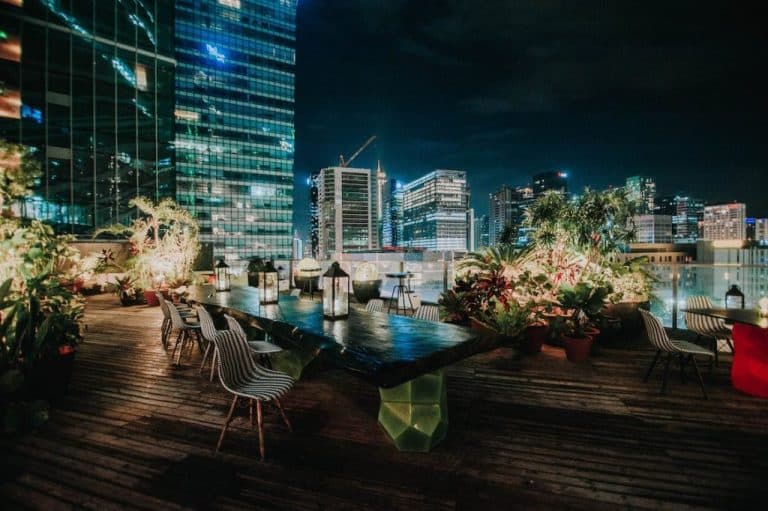 Bar rooftop noturno com mesa comunitária de madeira, cadeiras, lanternas e vasos de plantas ao ar livre com vista para prédios iluminados