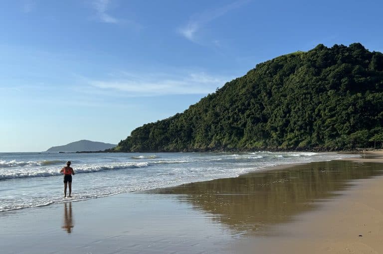 Pessoa de costas, usando chapéu e colete, em pé na beira d’água da Praia dos Amores, com pequenas ondas refletindo o céu azul e o Morro do Careca coberto pela Mata Atlântica ao fundo.